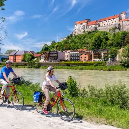 Zwei Radfahrer mit roten Rädern und blauen Taschen fahren auf einem Flussweg entlang der grünen Donau, mit einer Burg auf einem Hügel über einer Stadt.