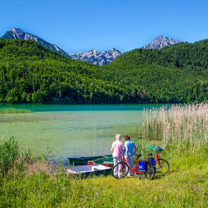 Zwei Radfahrer mit Rädern und Ruderboot am Ufer des Weißensees. Alpenberge und Wälder im Hintergrund unter blauem Himmel bei Füssen, Bayern.