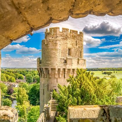 Blick auf den Steinturm von Warwick Castle durch einen verwitterten Bogen, mit grüner Landschaft und blauem Himmel dahinter.