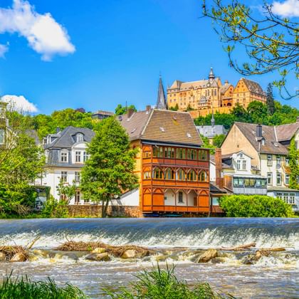 Marburger Landgrafenschloss auf Hügel über bunten historischen Gebäuden entlang der Lahn mit Wehr. Traditionelle Fachwerkhäuser zwischen grünen Bäumen.