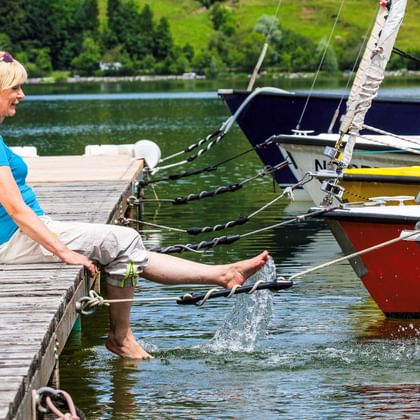 Frau in blauem Shirt sitzt auf Holzsteg mit Füßen im Wasser am Chiemsee. Bunte Segelboote liegen in der Nähe, grüne Hügel im Hintergrund.