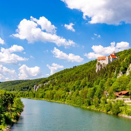 Burg Prunn thront auf weißen Kalksteinfelsen über der Altmühl, umgeben von grünen bewaldeten Hügeln unter blauem Himmel mit weißen Wolken.