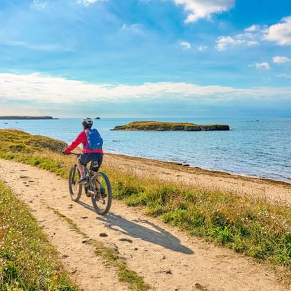 Radfahrerin in rosa Jacke fährt auf sandigem Küstenweg am Sillon du Talbert. Blaues Meer mit kleinen Inseln unter teilweise bewölktem Himmel.
