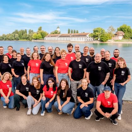 Große Gruppe von Radweg-Reisen Teammitgliedern in schwarzen und roten Shirts posiert an einem See mit historischen Gebäuden und Bäumen im Hintergrund.