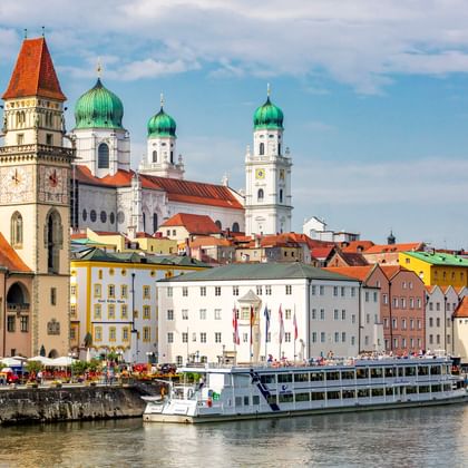 Passauer Altstadt mit bunten Gebäuden an der Donau. Ein Flusskreuzfahrtschiff liegt am Ufer. Kirchtürme mit grünen Kuppeln prägen die Skyline.
