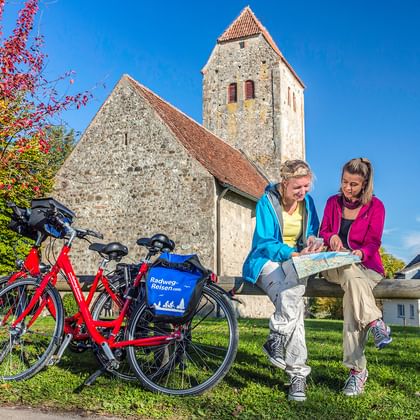 Zwei Radfahrerinnen studieren eine Karte während einer Pause bei einer historischen Steinkirche mit roten Fahrrädern auf Gras unter blauem Himmel.