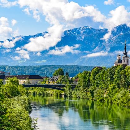 Fluss mit grünen Bäumen und Kirchtürmen, schneebedeckte Alpenberge im Hintergrund unter blauem Himmel mit weißen Wolken an der Alpe-Adria-Route.