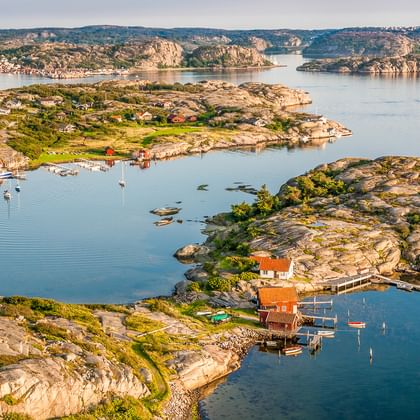 Luftaufnahme des schwedischen Westküsten-Schärengartens mit felsigen Inseln, roten Holzhäusern, Booten im ruhigen blauen Wasser und grüner Vegetation.