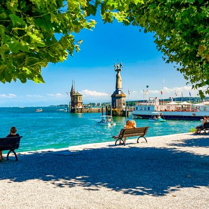 View through trees of Konstanz harbor on Lake Constance with the Imperia statue, lighthouse, and passenger ship. People sit on benches by the water.