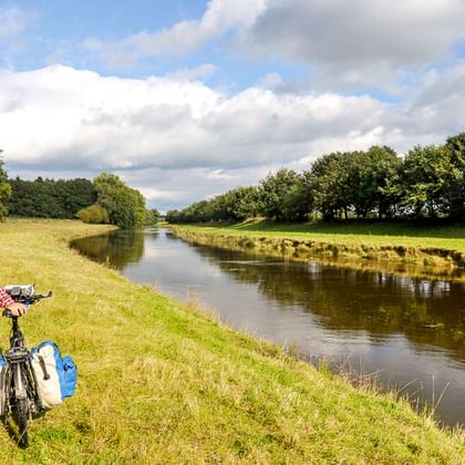 Radfahrer mit blauen Packtaschen steht am grasbewachsenen Flussufer neben der Ems. Grüne Wiesen und Bäume säumen beide Seiten unter bewölktem Himmel.
