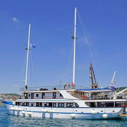 Weiße Motoryacht MS Aria mit blauem Zierstreifen und zwei Masten im Hafen. Weiße Fender entlang des Rumpfes, Küstenstadt im Hintergrund.