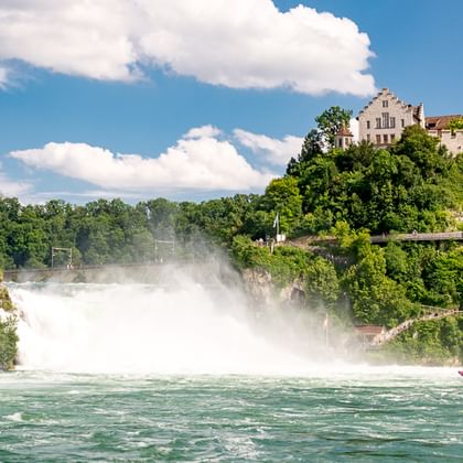 Rheinfall mit massiver Gischt und Schloss Laufen auf grünem Hügel. Ein rotes Touristenboot fährt im türkisfarbenen Wasser unterhalb des Falls.