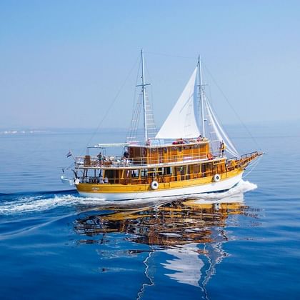 MS Planka, ein hölzernes Segelschiff mit weißen Segeln und zwei Masten, fährt auf ruhigem blauen Wasser mit fernen Bergen am Horizont.