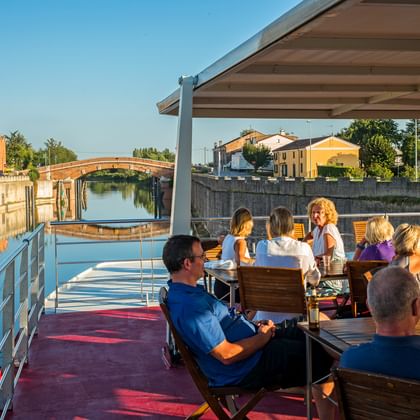 Passengers on the sun deck of MS Ave Maria in Portegrandi. View shows a canal with bridge, church tower, and Italian buildings under blue sky.