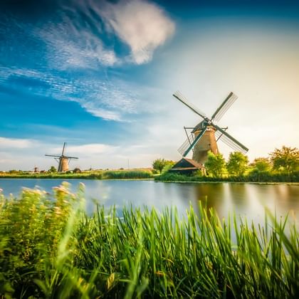 Traditionelle holländische Windmühle neben einem ruhigen Kanal mit grünem Schilf im Vordergrund. Blauer Himmel mit Schleierwolken.