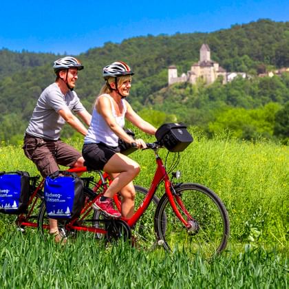 Zwei Radfahrer auf einem roten Tandemrad in einem grünen Feld mit Burg Kipfenberg auf einem bewaldeten Hügel im Hintergrund unter blauem Himmel.