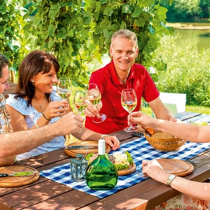 Vier Personen stoßen mit Weißwein an einem Tisch im Freien am Fluss an. Weinreben rahmen die Szene mit dem Main im Hintergrund ein.