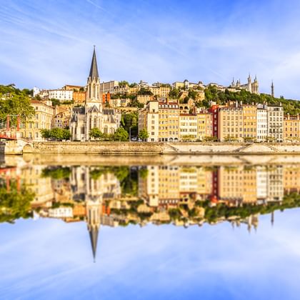Historic Lyon riverfront with colorful buildings and church spire perfectly mirrored in the calm waters of the Rhône River under blue sky.