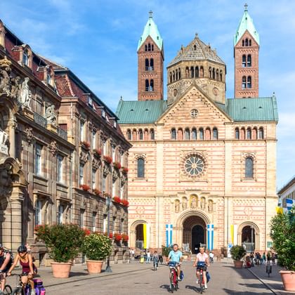 Radfahrer fahren über den Platz vor dem Speyerer Dom, einer romanischen Kirche mit Doppeltürmen und verzierter Fassade, eingerahmt von historischen Gebäuden.