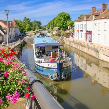 MS Fleur Flusskreuzfahrtschiff mit blauem Dach in einem schmalen Kanal in einem französischen Dorf, gesäumt von bunten Blumen und historischen Gebäuden.