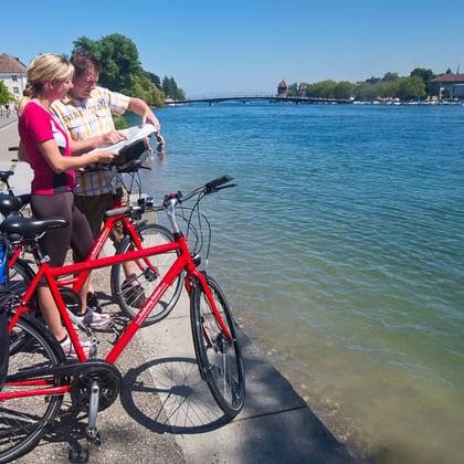 Paar mit Tourenrädern und blauen Packtaschen liest Karte an der Bodensee-Promenade. Klares blaues Wasser und Gebäude am gegenüberliegenden Ufer.