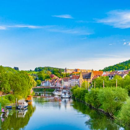 Malerischer Blick auf Wertheim mit dem Main, Booten am grünen Ufer, einem mittelalterlichen Steinturm und historischen Gebäuden unter blauem Himmel.