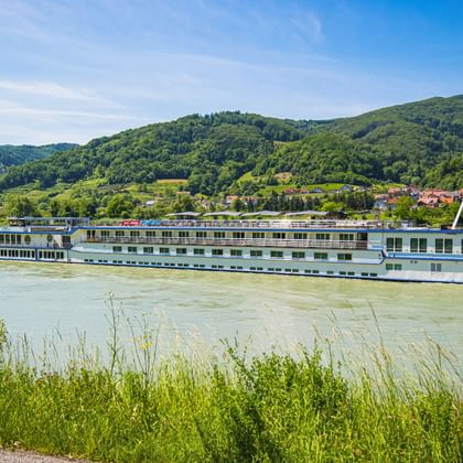 Weißes Flusskreuzfahrtschiff MS Swiss Crown auf der Donau mit grünen Hügeln, einem Dorf und blauem Himmel im Hintergrund.