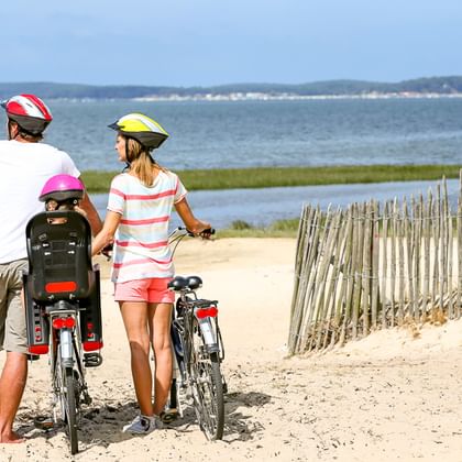 Drei Kinder mit Fahrrädern und Helmen auf sandigem Strandweg an der Ostsee, Blick auf Küstenlandschaft mit Wasser und ferner Küste.
