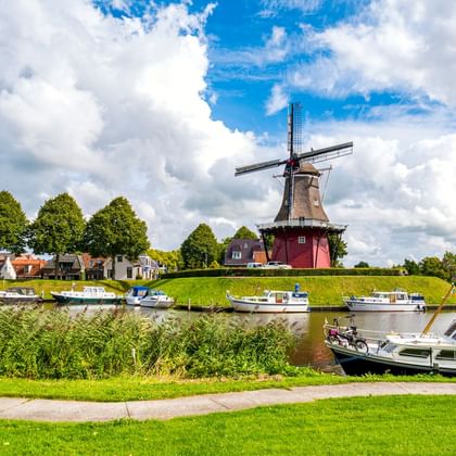 Traditionelle niederländische Windmühle auf Grasdamm in Dokkum mit Kanal, vertäuten Booten und Häusern unter blauem Himmel mit weißen Wolken.