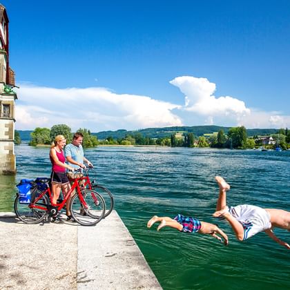 Zwei Radfahrer mit roten Rädern beobachten eine Person, die vom Uferweg in den Rhein springt. Historisches Gebäude und Hügel im Hintergrund.