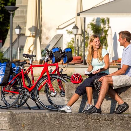 Zwei Radfahrer mit Tourenrädern sitzen auf Steinstufen beim Konzilgebäude in Konstanz und genießen eine Pause bei sonnigem Wetter.