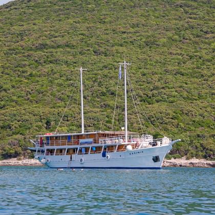 Weiße Motorsegelyacht MS Kazimir mit zwei Masten vor Anker in ruhigem blauen Wasser, mit dicht bewaldeter grüner Hügellandschaft im Hintergrund.