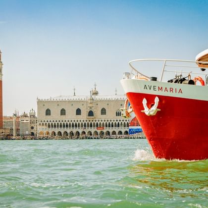 Rotweißes Schiff MS Ave Maria in der Lagune von Venedig mit Markusturm und Dogenpalast im Hintergrund unter blauem Himmel.