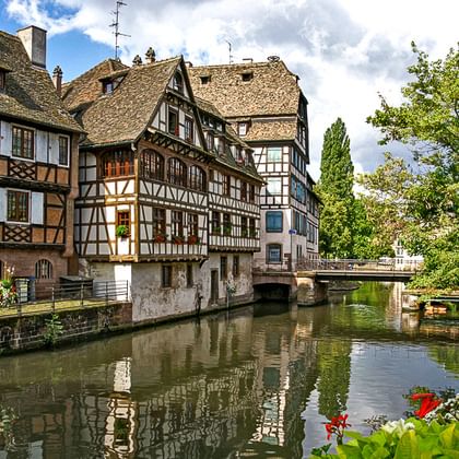 Traditionelle Fachwerkhäuser säumen einen ruhigen Kanal im Elsass. Eine Steinbrücke überquert das Wasser, bunte Blumen im Vordergrund.