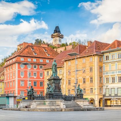 Hauptplatz square in Graz with ornate fountain monument in center, colorful historic buildings with red-tiled roofs, and iconic clock tower on hill.