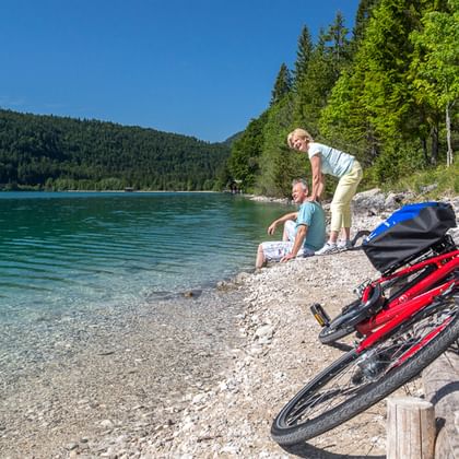 Zwei Radfahrer rasten an einem Kiesstrand neben einem türkisfarbenen See in Oberbayern. Zwei rote Fahrräder mit Taschen stehen im Vordergrund.