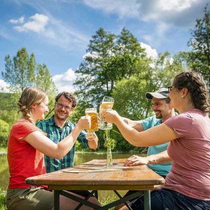 Vier Radfahrer stoßen mit Biergläsern an einem Holztisch an der Altmühl an, umgeben von grünen Bäumen unter blauem Himmel.