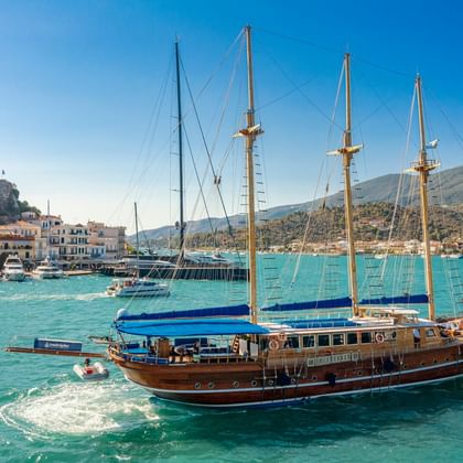 Hölzernes Segelschiff MS Thalassa mit vier Masten in türkisfarbenem Wasser nahe der Insel Poros, mit weißen Gebäuden und Hügeln im Hintergrund.