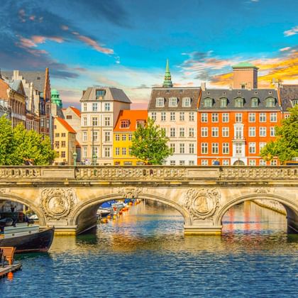 Steinbrücke mit verzierten Bögen über einem Kanal in Kopenhagens Altstadt, gesäumt von bunten historischen Gebäuden unter dramatischem Abendhimmel.