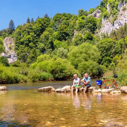 Zwei Radfahrer kühlen ihre Füße in der Donau bei Fridingen, mit roten Tourenrädern am felsigen Ufer und Kalksteinfelsen im Hintergrund.