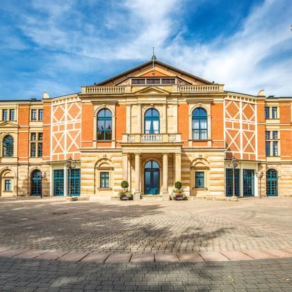 The Bayreuth Festspielhaus, a grand orange and cream-colored opera house with arched windows and decorative patterns, framed by green trees.