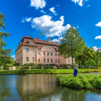 Schloss Branitz in Cottbus, ein beiges Barockgebäude mit rotem Dach, spiegelt sich in einem Teich. Üppige grüne Rasenflächen und Bäume umgeben das Schloss.