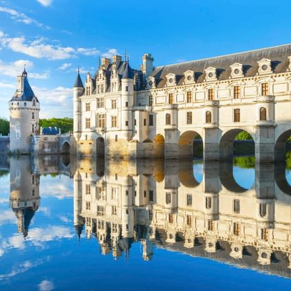 Château de Chenonceau in Loire Valley with white Renaissance facade and arched bridge spanning calm water, creating perfect mirror reflection under blue sky.