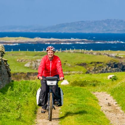 Radfahrer in roter Jacke mit Tourenrad auf grasigem Küstenweg in Connemara. Steinruinen und blaues Meer mit Bergen im Hintergrund.