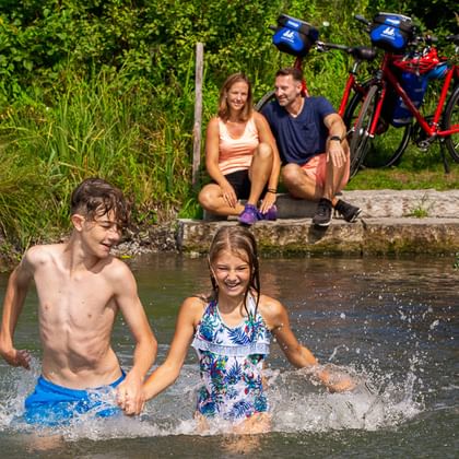 Two children playing in a stream while parents sit on the bank with bicycles in the background at Lake Constance.