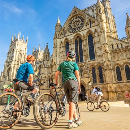 Zwei Radfahrer mit Rädern vor der Kathedrale York Minster in Yorkshire. Die gotische Kathedrale zeigt eine große Rosette und ornamentale Fassade.