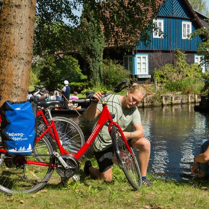 Zwei Radfahrer mit roten Rädern rasten an einem Kanal im Spreewald. Blaues traditionelles Haus am Wasser, umgeben von grünen Bäumen.