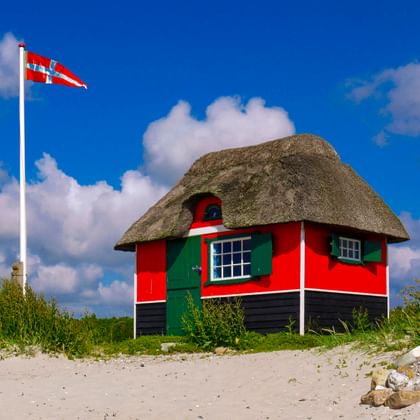 Rot-schwarze Strandhütte mit Reetdach und grünen Fensterläden am Sandstrand. Dänische Flagge weht an weißem Mast unter blauem Himmel mit Wolken.