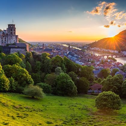 Heidelberger Schloss auf einem Hügel bei Sonnenuntergang mit Blick auf das Neckartal, grünen Wiesen im Vordergrund und historischer Stadt.
