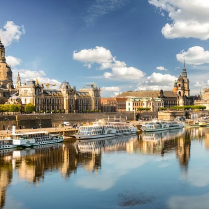 Dresdens historische Skyline entlang der Elbe mit barocker Architektur und Kuppelkirchen. Mehrere Flusskreuzfahrtschiffe liegen am Ufer.